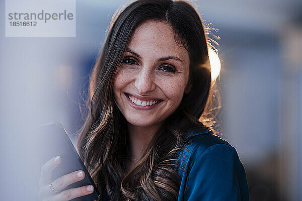 Glückliche junge Frau mit braunen Haaren hält Smartphone in der Hand