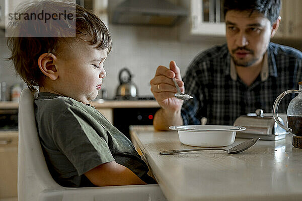 Vater füttert verärgerten Sohn  der zu Hause am Esstisch sitzt