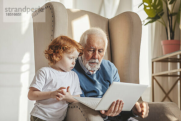 Großvater teilt Laptop und sitzt zu Hause im Sessel