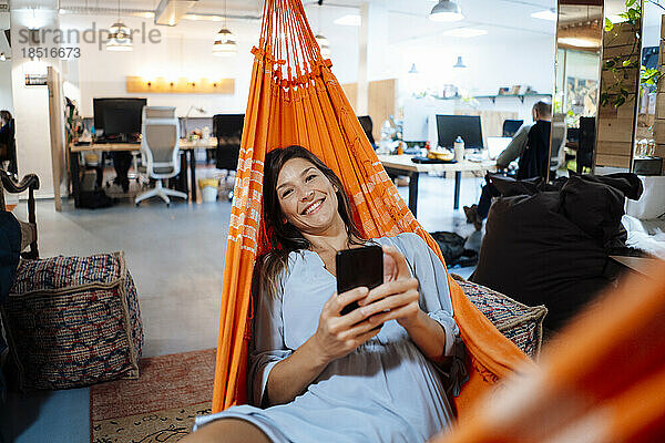Glückliche Geschäftsfrau hält Smartphone in der Hängematte im Büro liegend