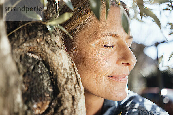 Reife Frau mit geschlossenen Augen umarmt Baum an sonnigem Tag