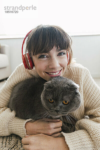 Glückliche Frau mit kabellosen Kopfhörern umarmt Katze zu Hause