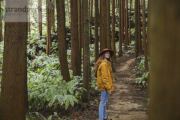 Rothaarige Frau mit Hut steht inmitten hoher Bäume im Wald
