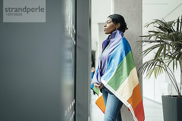 Junge Frau mit Regenbogenfahne bedeckt  die aus dem Fenster im Büro schaut