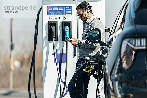 Mann benutzt Smartphone und stützt sich am Bahnhof auf Elektroauto