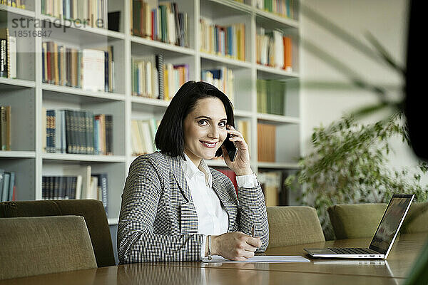 Lächelnde Geschäftsfrau  die am Schreibtisch im Büro sitzt und mit einem Smartphone spricht