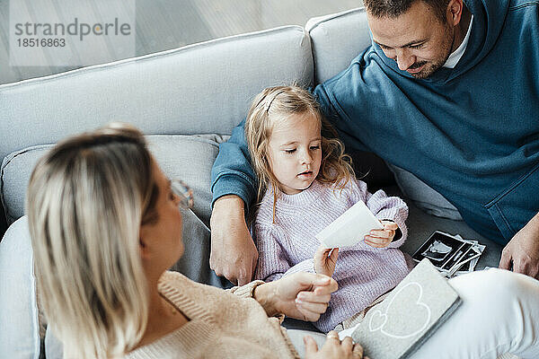 Tochter hält Ultraschallbild von Eltern  die auf Sofa im Wohnzimmer sitzen