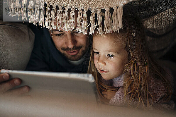 Father sharing tablet PC with daughter under blanket at home
