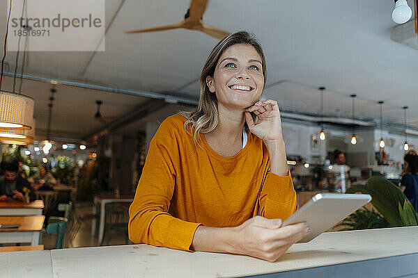 Glückliche Frau hält Tablet-PC und sitzt mit der Hand am Kinn im Café