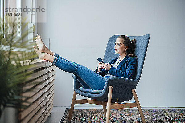 Glückliche junge Geschäftsfrau hält Smartphone im Sessel sitzend