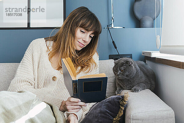 Frau zeigt der Katze  die zu Hause auf dem Sofa sitzt  ein Buch