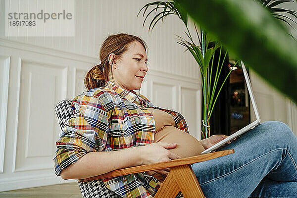 Lächelnde schwangere Frau  die zu Hause mit Laptop auf einem Stuhl sitzt