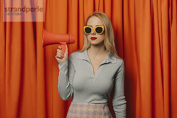 Frau mit Sonnenbrille hält Megafon vor orangefarbenem Vorhang