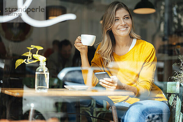 Glückliche Frau  die mit einer Kaffeetasse im Café sitzt und durch Glas gesehen wird