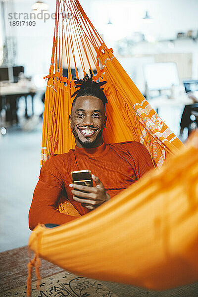 Glücklicher junger Geschäftsmann entspannt sich in der Hängematte mit Smartphone im Büro