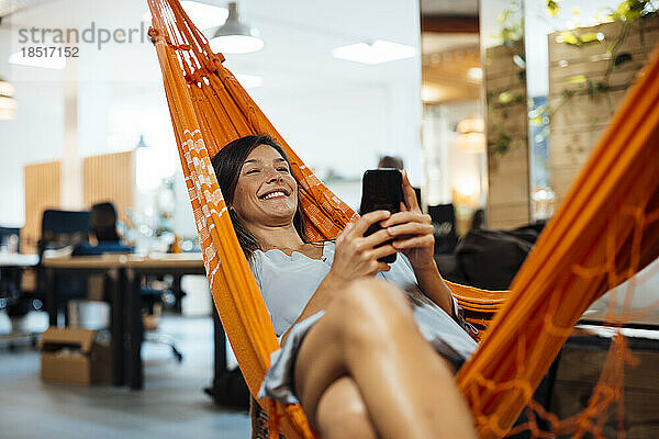 Lächelnde Geschäftsfrau benutzt Smartphone und entspannt sich in der Hängematte im Büro