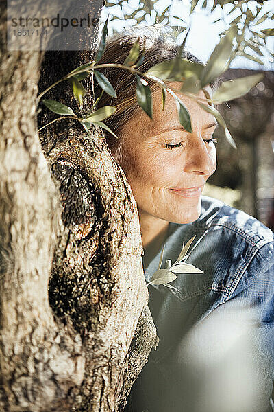 Lächelnde reife Frau mit geschlossenen Augen umarmt Baum an sonnigem Tag