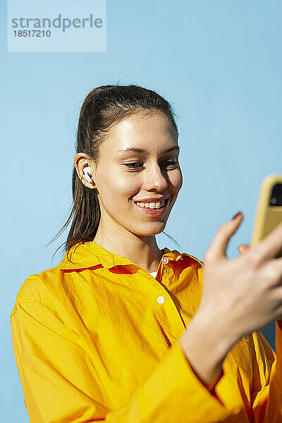 Glückliche Frau mit kabellosen In-Ear-Kopfhörern und Mobiltelefon vor blauem Hintergrund