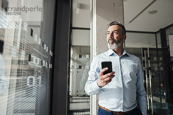 Nachdenklicher Geschäftsmann steht mit Smartphone im Büro