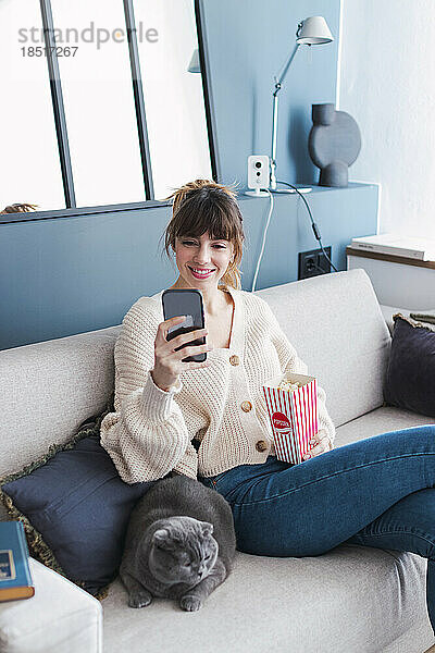 Lächelnde Frau mit Popcorn und Smartphone  die zu Hause auf dem Sofa sitzt