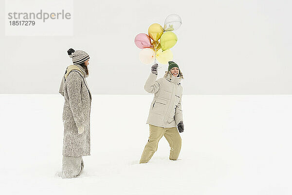 Lächelnde Frau mit glänzenden Luftballons  die Spaß mit einer Freundin im Schnee hat