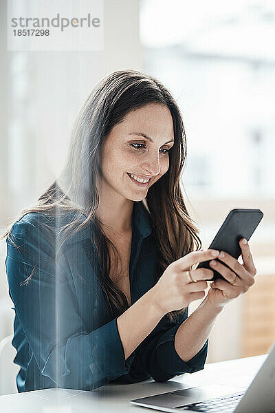 Glückliche junge Geschäftsfrau  die im Heimbüro ihr Smartphone nutzt