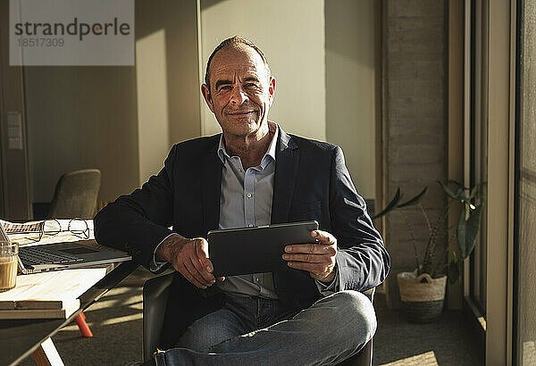 Lächelnder reifer Geschäftsmann sitzt mit Tablet-PC im Büro