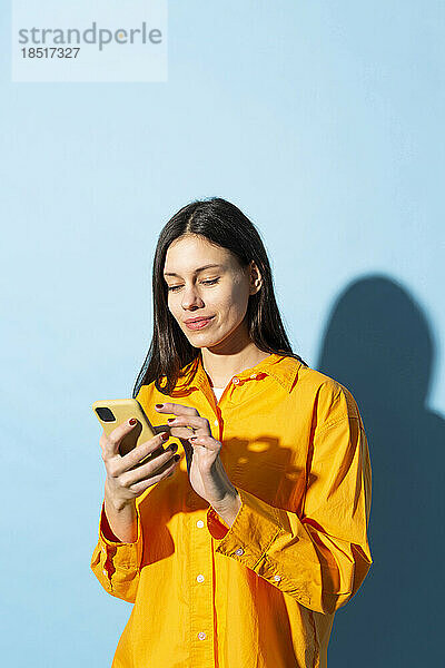 Lächelnde junge Frau  die ihr Mobiltelefon vor blauem Hintergrund benutzt