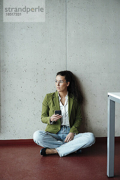 Nachdenkliche junge Geschäftsfrau mit Smartphone sitzt auf dem Boden im Büro
