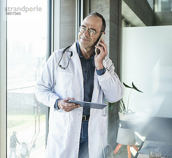 Reifer Arzt mit Tablet-PC  der am Fenster auf dem Smartphone spricht