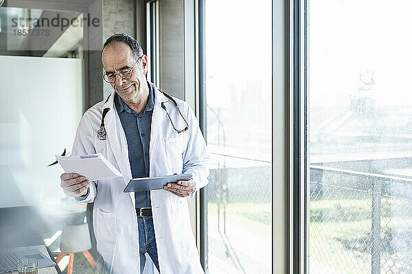 Lächelnder reifer Arzt liest Rezept und hält Tablet-PC am Fenster