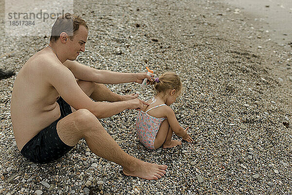 Mann trägt Sonnencreme auf Tochter auf  die am Strand spielt