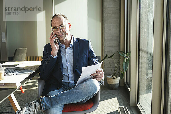 Reifer Geschäftsmann spricht am Smartphone und hält ein Dokument im Büro