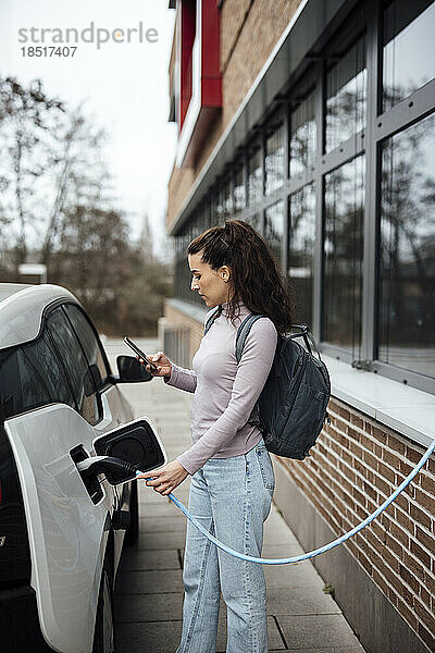 Lächelnde Frau benutzt Smartphone und lädt Elektroauto auf