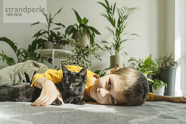 Lächelnder Junge liegt mit schwarzer Katze auf dem Teppich
