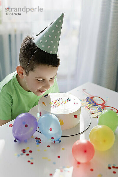 Lächelnder Junge blickt inmitten von Luftballons und Konfetti auf die Geburtstagstorte