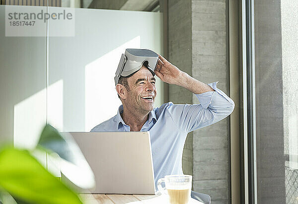 Glücklicher reifer Geschäftsmann mit Virtual-Reality-Simulator sitzt am Schreibtisch im Büro