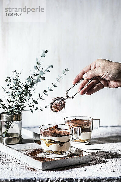 Hand einer Frau streut Kakaopulver durch ein Sieb auf Tiramisu im Becher