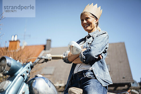 Glückliche reife Frau mit Krone  die mit verschränkten Armen auf dem Motorrad steht