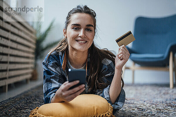Lächelnde Geschäftsfrau mit Smartphone und Kreditkarte auf Teppich liegend
