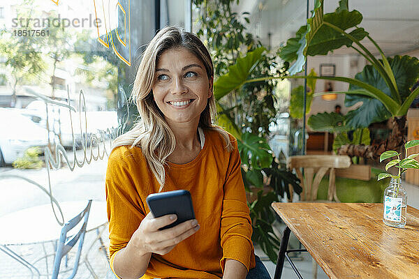 Glückliche Frau mit Smartphone  die durch das Fenster im Café schaut