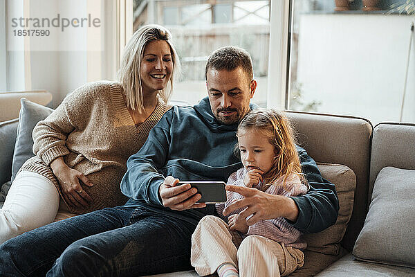 Parents sharing smart phone with daughter sitting on sofa at home