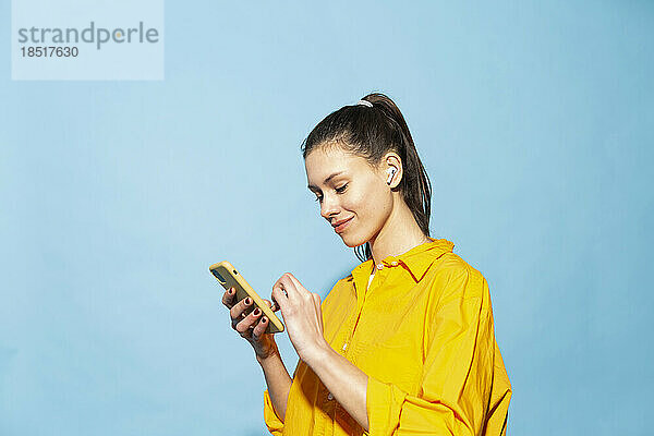 Lächelnde junge Frau  die mit ihrem Mobiltelefon vor blauem Hintergrund im Internet surft