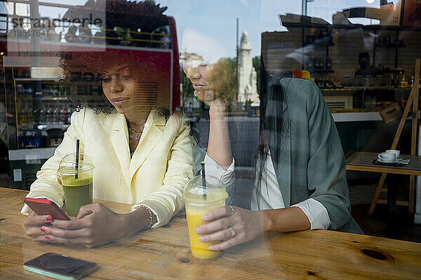 Frau teilt Smartphone mit Freundin  die im Café sitzt und durch das Fenster gesehen wird