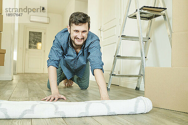 Glücklicher junger Mann rollt Teppich im neuen Zuhause aus