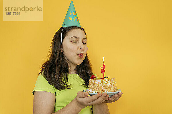 Mädchen bläst Kerze auf Geburtstagstorte vor gelbem Hintergrund