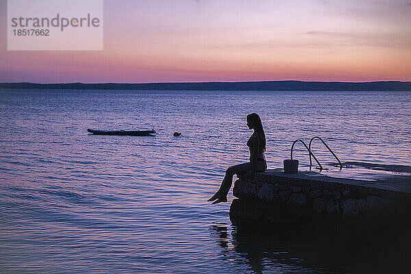 Silhouette einer Frau  die bei Sonnenuntergang auf einem Steg am Meer sitzt