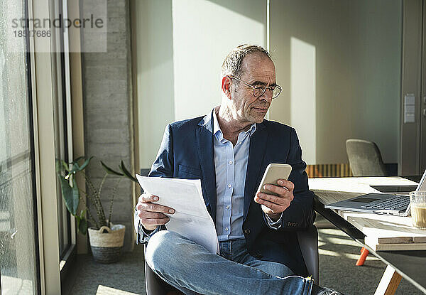 Reifer Geschäftsmann nutzt Smartphone und sitzt mit Dokument im Büro