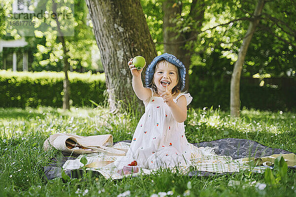 Fröhliches Mädchen gestikuliert und zeigt grünen Apfel im Garten