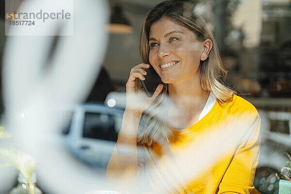 Glückliche Frau  die im Café ihr Smartphone nimmt und durch Glas gesehen wird